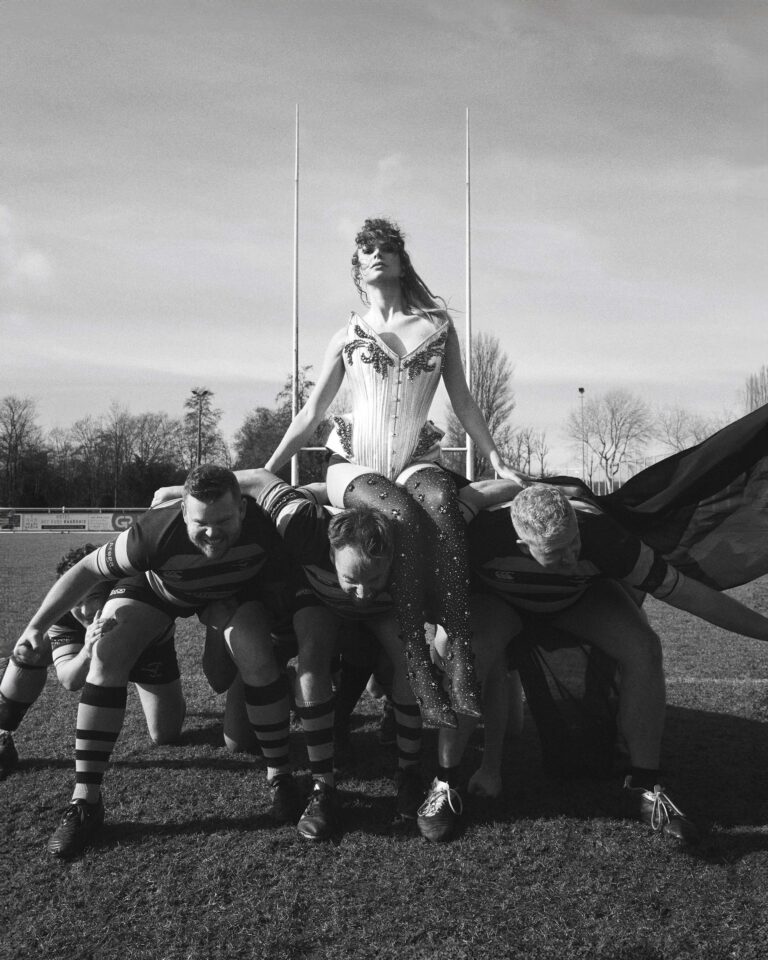 Zwart-wit afbeelding van een centrale figuur met lange cape die rechtop staat, terwijl drie mensen ernaast neerknielen.