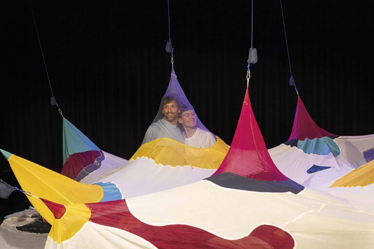 Veelkleurige, hangende doeken in tentachtige vormen tegen een donkere achtergrond.