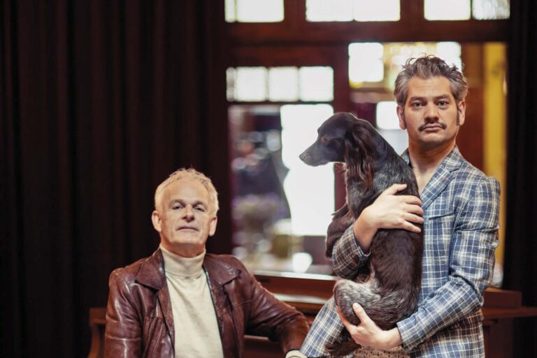 Twee mannen poseren in een kamer, met een zittende man in een rode leren jas en een staande man met blond haar.