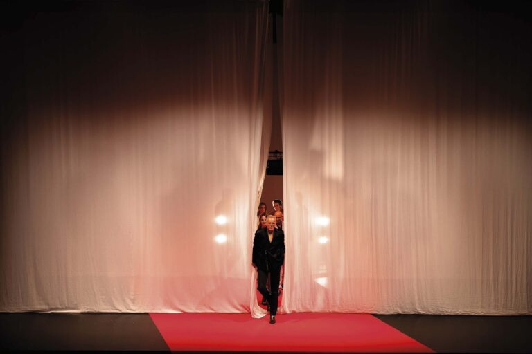 Een persoon loopt over een rode loper op een podium, met gordijnen op de achtergrond en felle lichten.