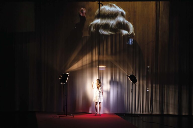 Een persoon staat in het midden van een donker podium, verlicht door een spotlight, met een rode loper en een houten achterwand op de achtergrond.