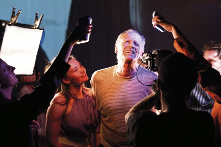 Een groep mensen heft hun drankjes omhoog terwijl een glimlachende man in het midden onder kleurrijk podiumlicht staat.