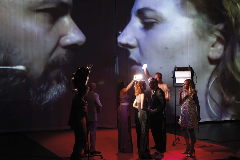 Een tentoonstellingsruimte met een grote projectie van gezichten op de muur, terwijl bezoekers eromheen staan.