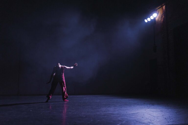 Een danser op een donker podium onder blauw-paars licht, met een uitgestrekte arm.