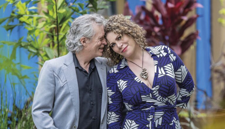 Een paar staat dicht bij elkaar en glimlacht buiten; de vrouw draagt een blauwe jurk met witte patronen en de man een lichtgrijze blazer.