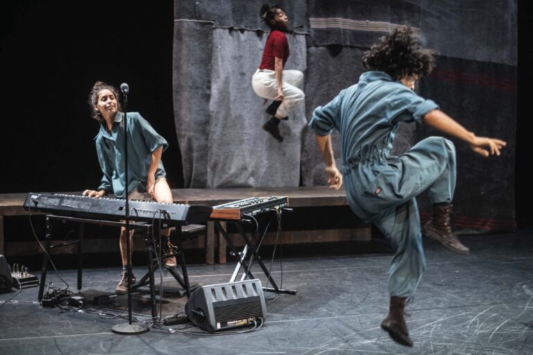 Een danser maakt een sprong op een podium terwijl een muzikant achter een keyboard speelt.