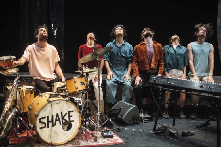 Een band op een podium met een drumstel waarop de tekst “SHAKE” staat.