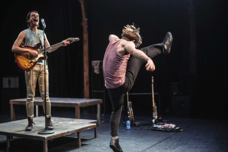 Een danser op een donker toneel doet een stretch met één been omhoog tegen een houten paal, terwijl iemand in de achtergrond toekijkt.