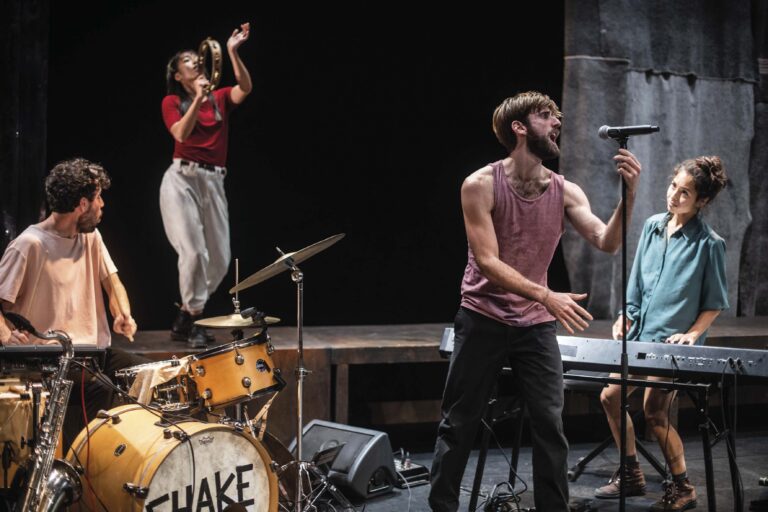 Twee muzikanten geven een live optreden op een donker podium, met een drumstel op de achtergrond.