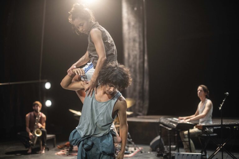Een danser in een blauwe outfit danst op een donker podium, met andere dansers en felle podiumlichten op de achtergrond.