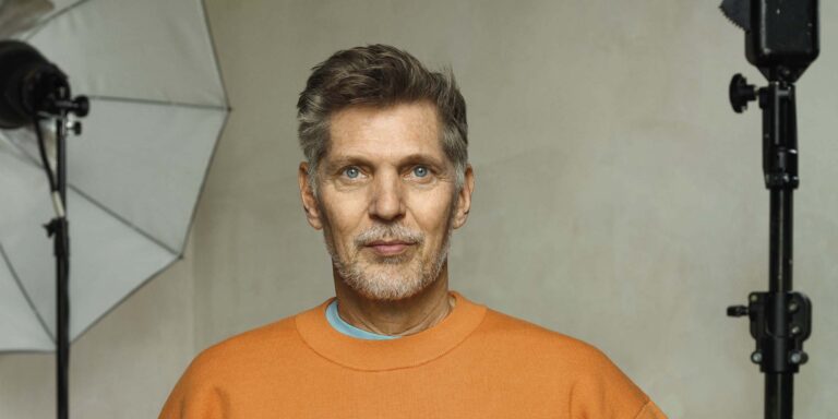 Een man met korte grijze haren en een oranje trui, kijkend naar de camera tegen een neutrale achtergrond.