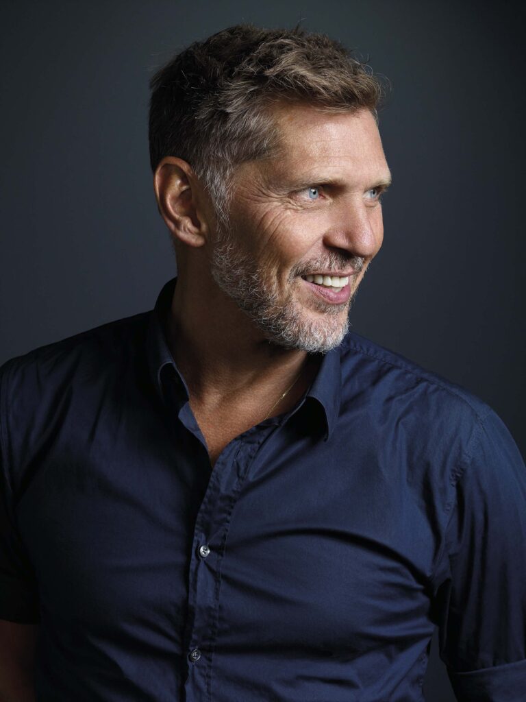 Glimlachend portret van een man met kort grijs haar en een donkerblauw overhemd tegen een donkere achtergrond.