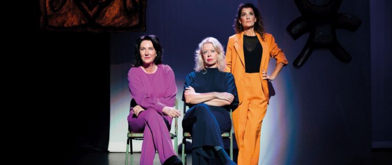 Drie vrouwen in modieuze outfits zitten op stoelen, met verschillende gezichtsuitdrukkingen en posities.
