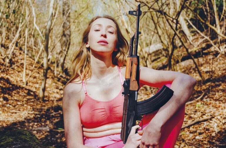 Vrouw in een roze top staat in een bos en houdt een geweer vast.