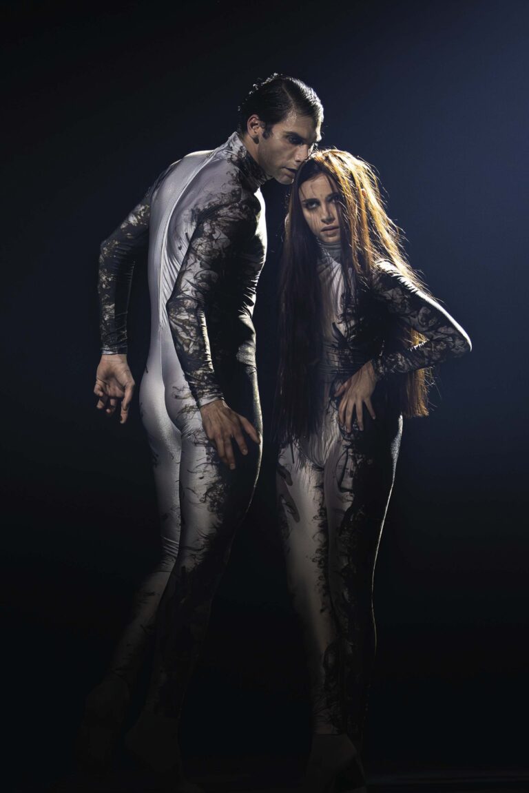 Twee modellen in glanzende zilveren outfits poseren tegen een donkere achtergrond.