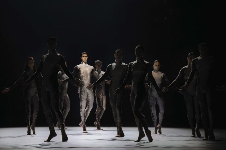 Een groep dansers op een donker podium in donkere kleding tijdens een dansvoorstelling.