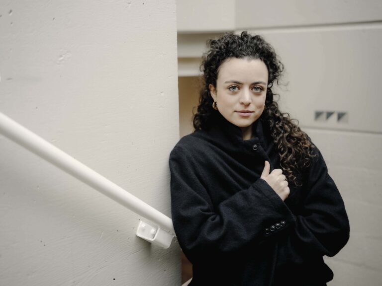 Een vrouw met donker krullend haar staat bij een witte trapleuning en kijkt naar de camera.