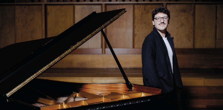 Een vleugelpiano met het deksel omhoog en een man in een donker pak staand naast de piano.