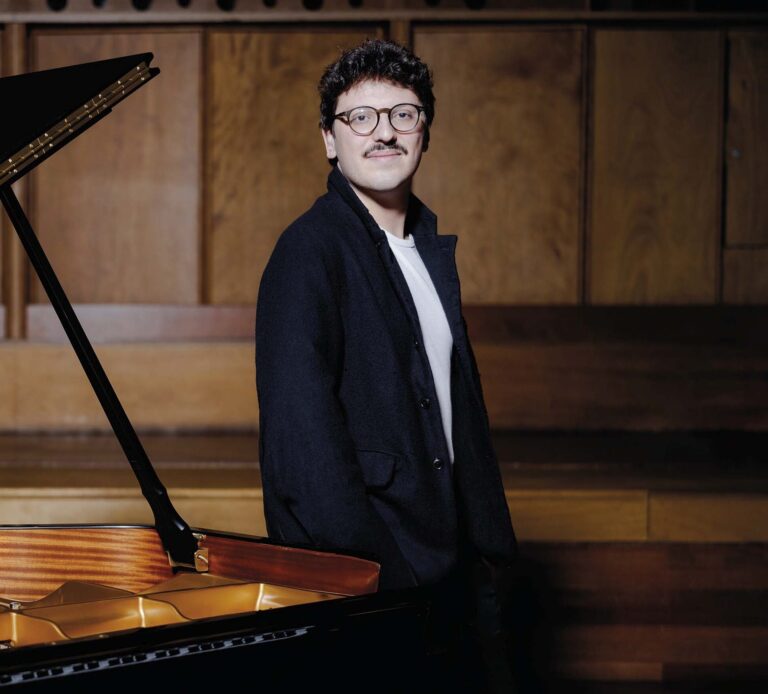 Een vleugelpiano met het deksel omhoog en een man in een donker pak staand naast de piano.