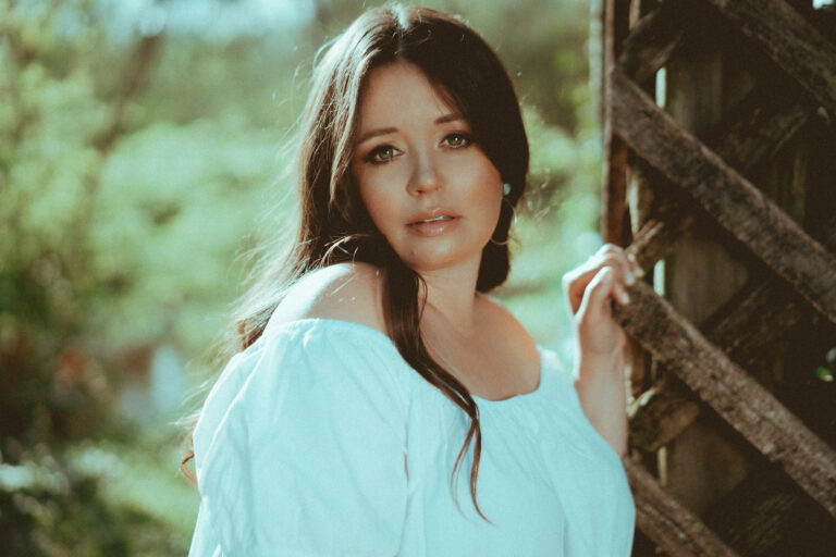 Een vrouw met lang, donker haar poseert vriendelijk in een groene, natuurlijke omgeving.