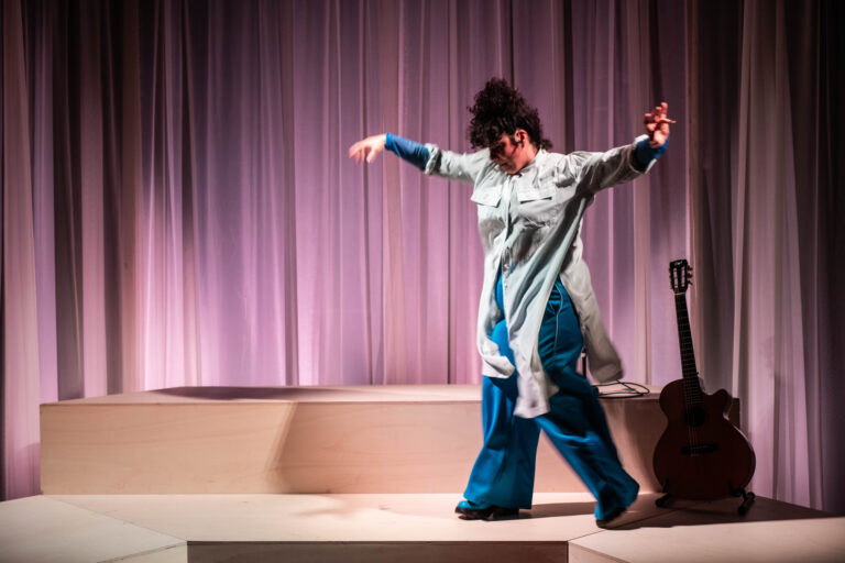 Een danseres in losse kleding beweegt zich elegant op een podium met een gitaar op de achtergrond.