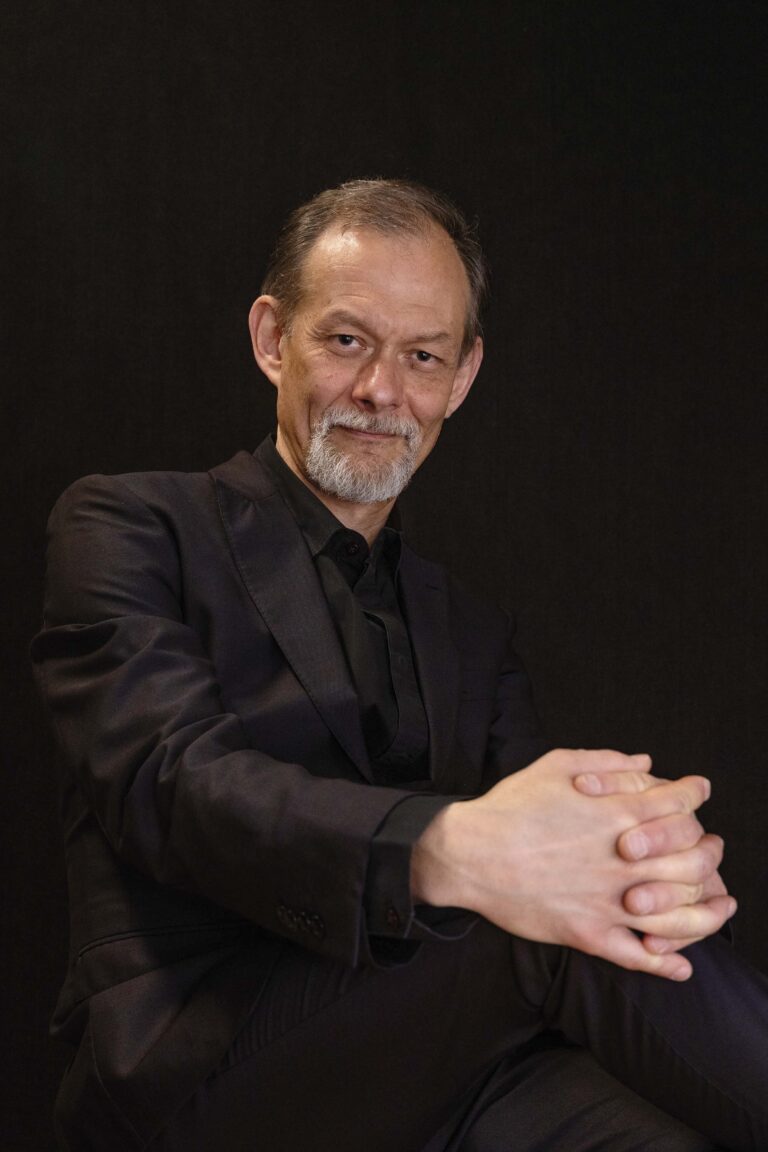 Oudere man met grijze baard in een donker pak tegen een donkere achtergrond, met zijn handen voor zich gevouwen.