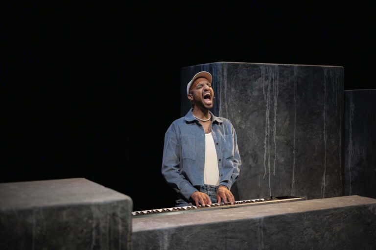 Een man in een spijkerjasje staat achter een houten lessenaar op een donker podium en zingt.