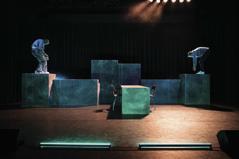 Op een donker toneel staat een persoon tussen grote blauwgroene blokken als decor.