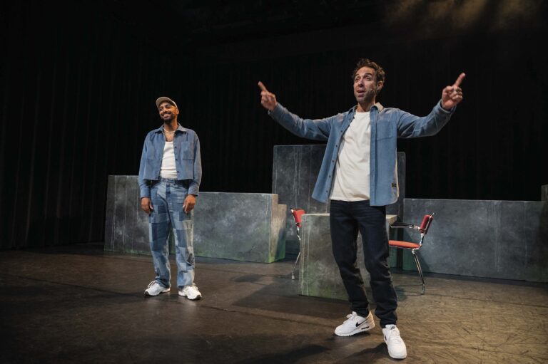 Twee mannen in casual kleding staan op een donker theaterpodium tijdens een voorstelling, met één van hen armen wijd.