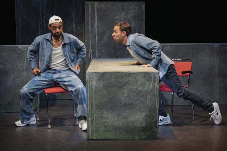 Twee mensen in denim outfits staan naast een grote grijze kubus op een donker toneel.