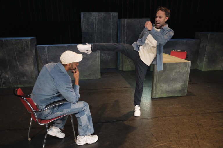 Twee mannen op een donker theaterpodium, een zittende man met een witte hoed en hoofd gebogen terwijl de staande man naar hem wijst.