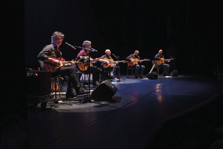 Een groep gitaristen zit op een donker podium onder blauwe belichting tijdens een live optreden.