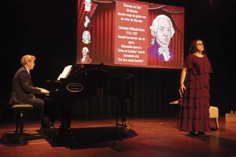 Podium met een vleugel links en een spreker rechts, voor een grote schermpresentatie met een portret en tekst op een rode achtergrond.