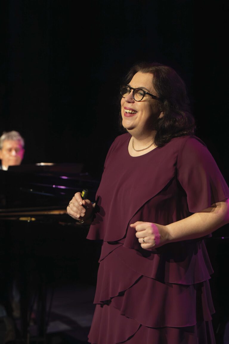 Een vrouw in een paarse jurk zingt op een podium, met een pianist op de achtergrond.
