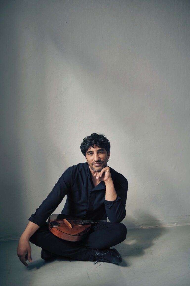 Een man met donker haar zit tegen een lichte muur en steunt zijn kin op zijn hand.