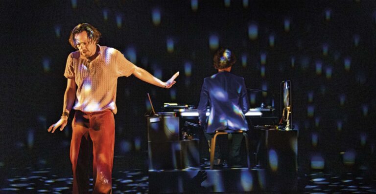 Twee artiesten op een verlicht podium, terwijl een van hen bij een mengpaneel staat.