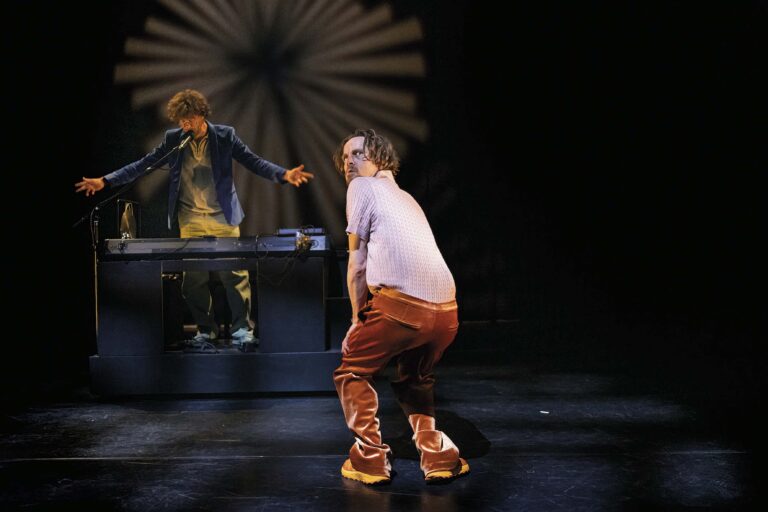 Een danser in oranje broek en witte shirt danst op een podium terwijl een DJ achter een mengpaneel op de achtergrond staat.