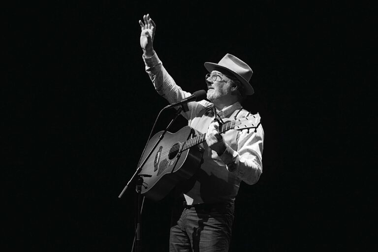 Zwart-wit foto van een muzikant met hoed die op een podium gitaar speelt en achter een microfoon zingt.