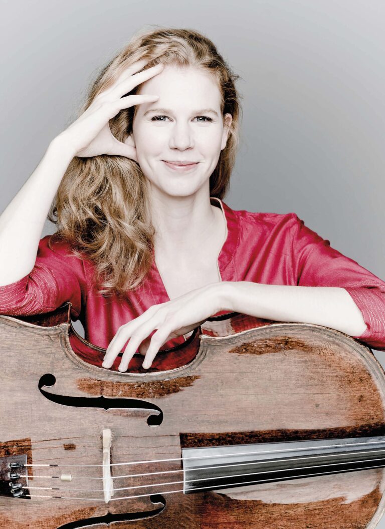 Vrouw met rode blouse houdt een cello vast en glimlacht naar de camera.