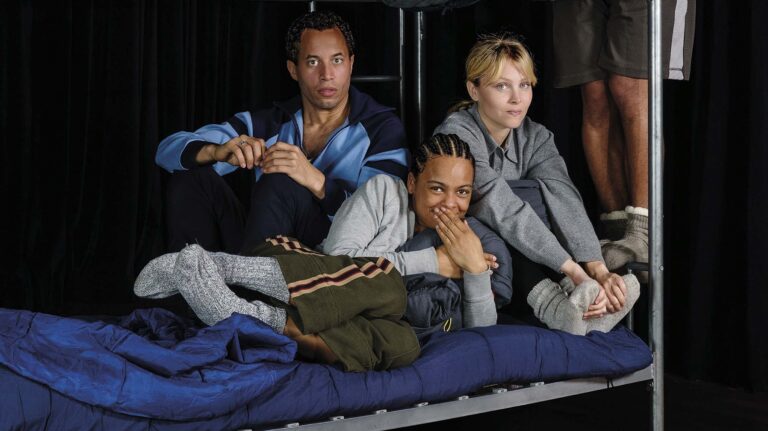 Drie jonge mensen zitten samen op een bank met een blauw dekentje en kijken naar de camera.