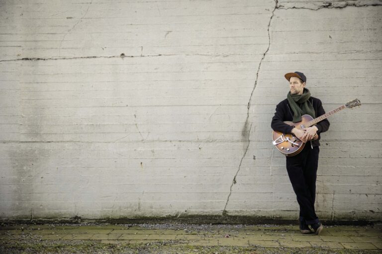 Een man met een gitaar staat tegen een witte muur met een zichtbare scheur.