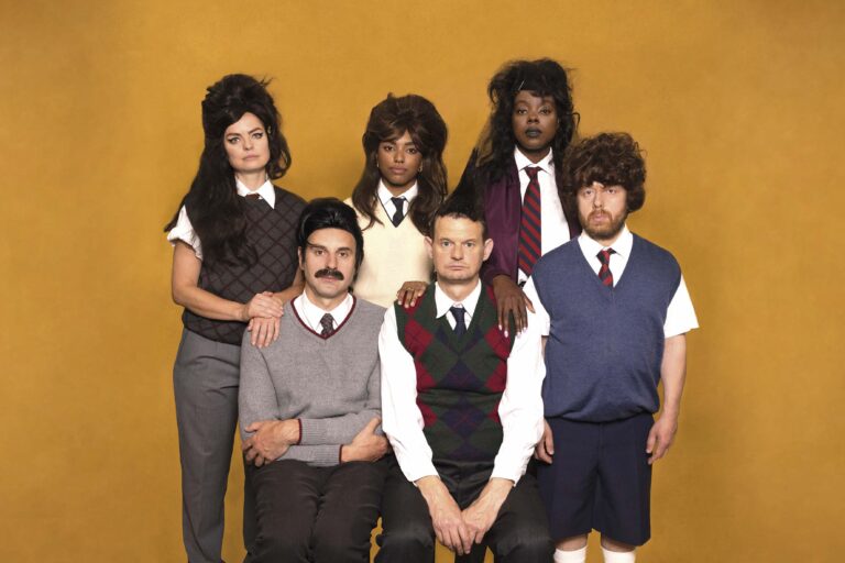 Zes personen poseren in twee rijen voor een groepsportret, gekleed in opvallende jaren-70-stijl outfits.