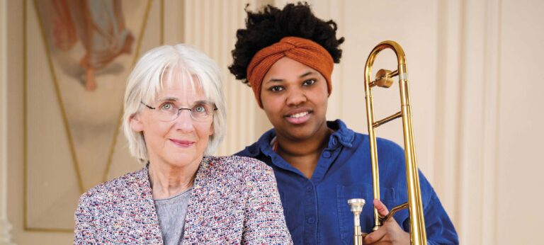 Een oudere vrouw en een jonge tromboniste staan naast elkaar en glimlachen, met een trombone zichtbaar.