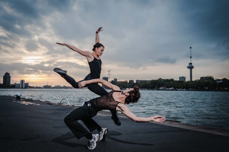 Twee dansers voeren boven het water een acrobatische sprong uit, met de stadsachtergrond op de achtergrond.