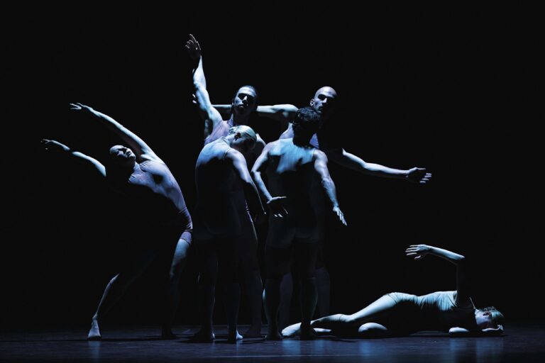 Meerdere balletdansers bewegen op een donker podium, verlicht door koel blauw licht.