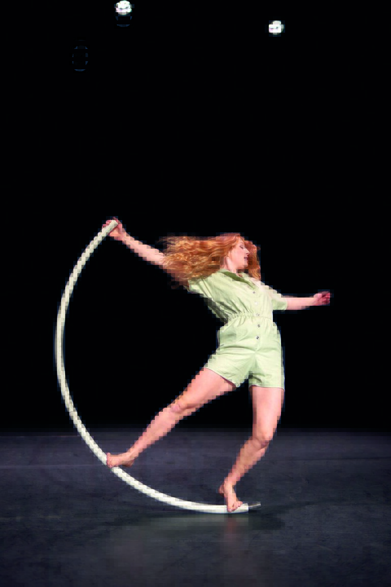 Een acrobate met lange, golvende haren voert een truc uit met een hula hoop op een donkere achtergrond.