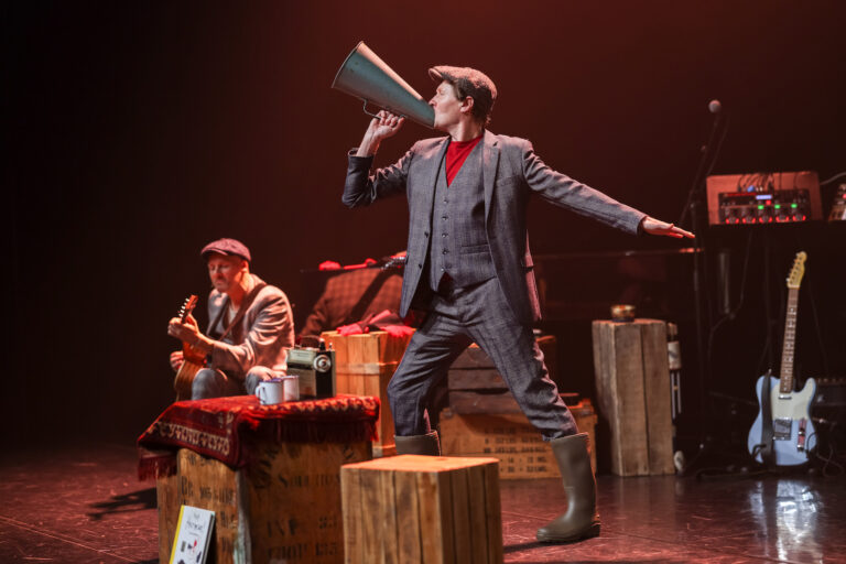 Een acteur in een gestreept pak staat op een kist met een megfoon, terwijl een muzikant op de achtergrond gitaar speelt.
