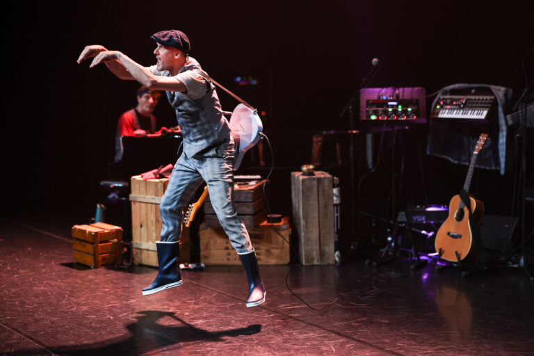 Een performer in een opvallend kostuum danst energiek op een podium, omringd door muziekinstrumenten en houten kisten.
