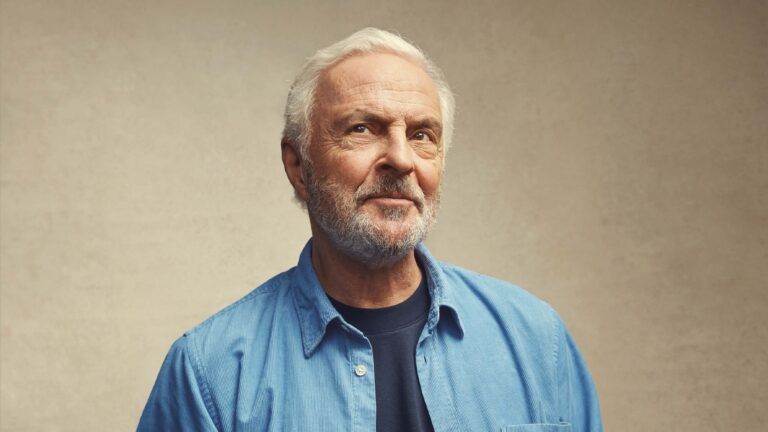 Portret van een oudere man met grijs haar en een baard, die een blauw overhemd draagt tegen een beige achtergrond.
