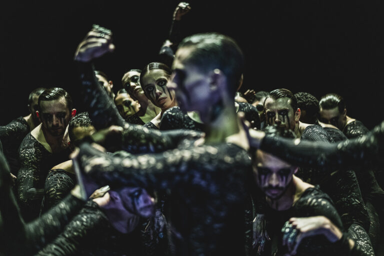 Een groep dansers met een donkere, glimmende huid, die een intense pose aanneemt op een donkere achtergrond.
